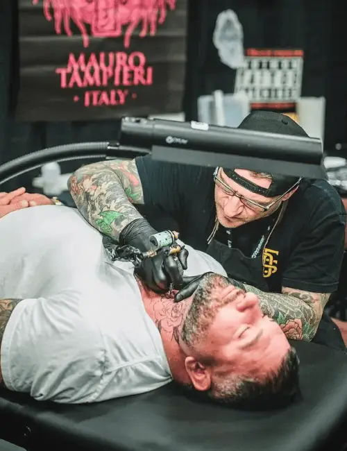 Un tatuatore di Tatuaggi Giapponesi con i guanti sta tatuando il collo di un uomo sdraiato su un tavolo. Entrambi hanno tatuaggi visibili. In questo studio di tatuaggi di Ravenna sono presenti attrezzature per tatuaggi e un'insegna.